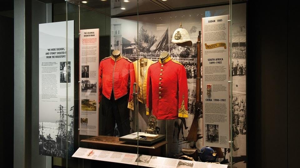 A colonial-era display case of red tunics, a pith helmet and a rifle against panels on Sudan 1885