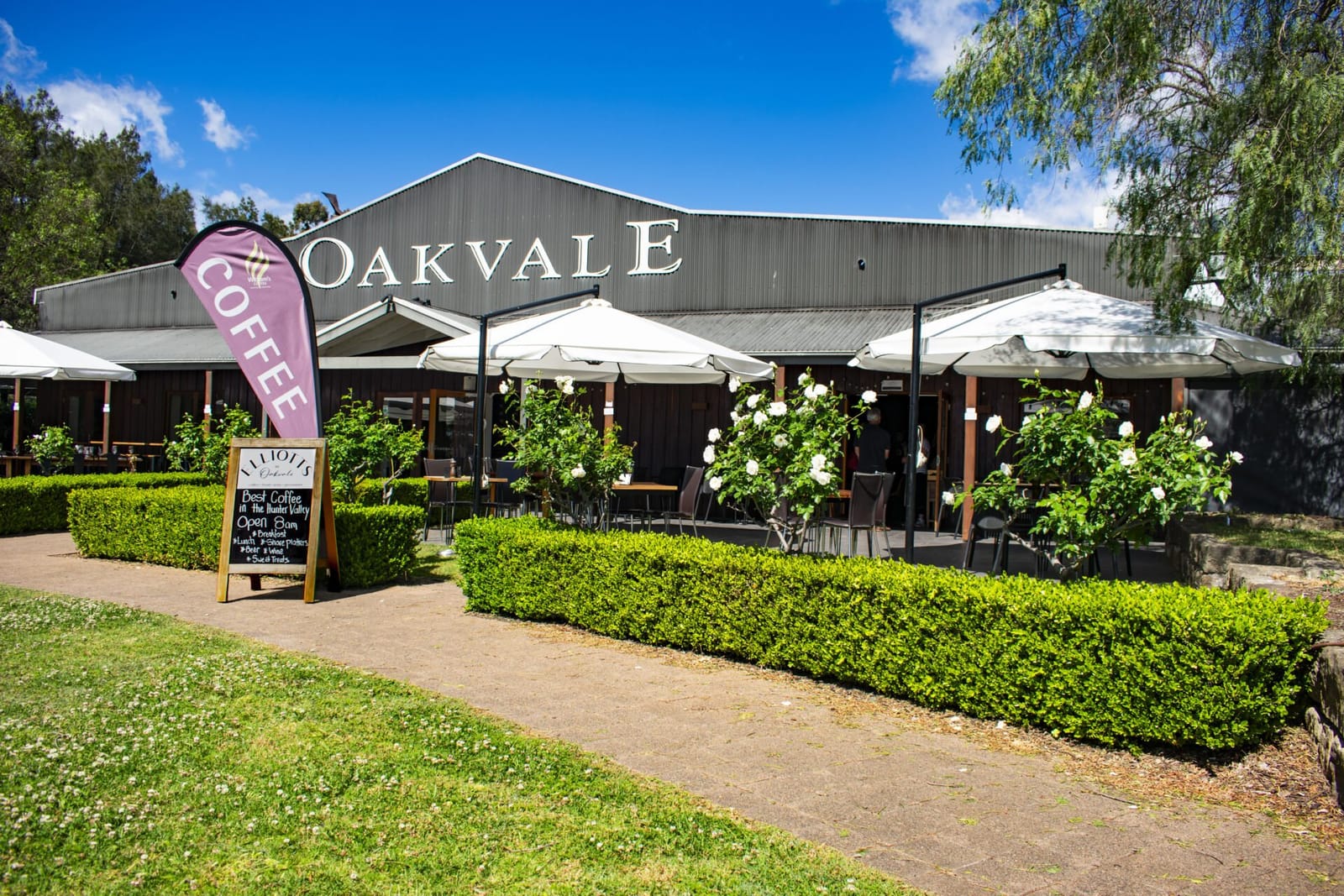 Elliotts at Oakvale exterior with the corrugated-iron Oakvale building, umbrella-shaded tables and a coffee flag out front.
