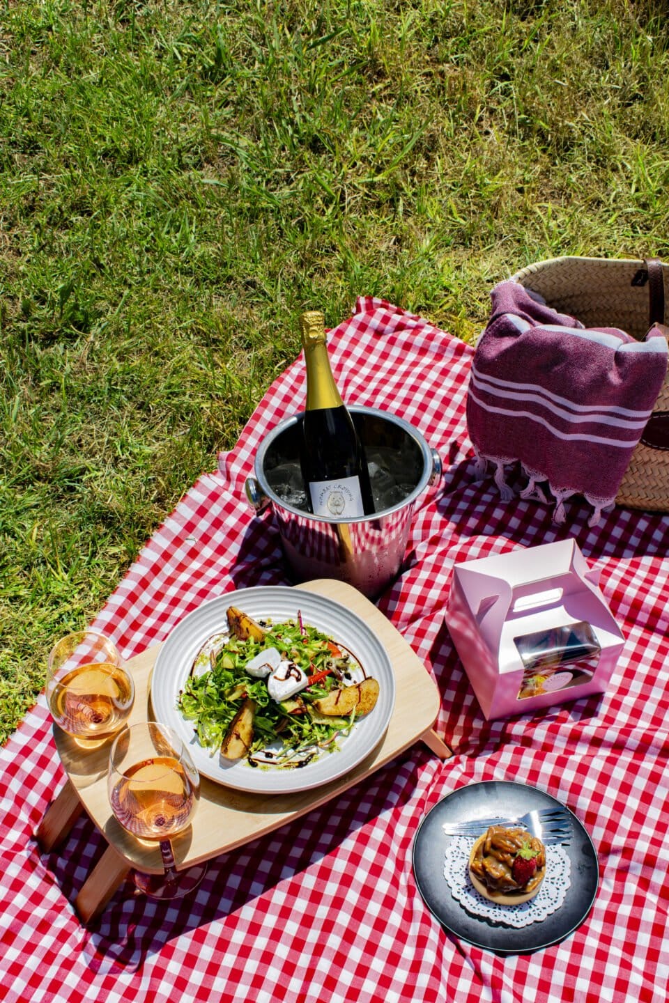 A picnic spread of salad, a plated dessert and chilled sparkling wine on a checked blanket at Elliotts at Oakvale.