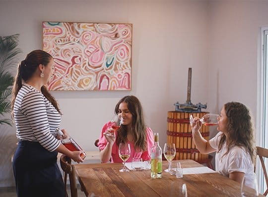 Three women in tasting room with wine service at wooden table
