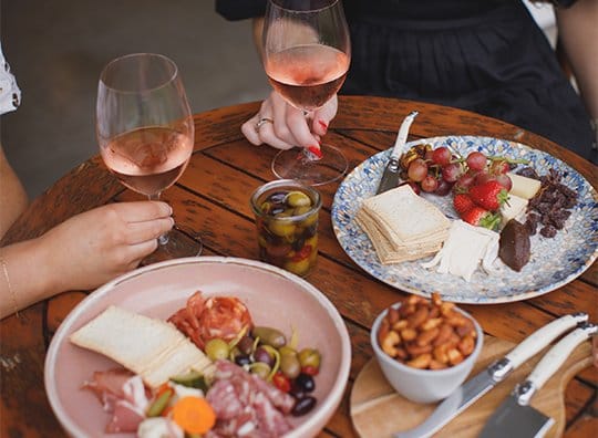 Hands holding rose wine glasses with charcuterie board nearby
