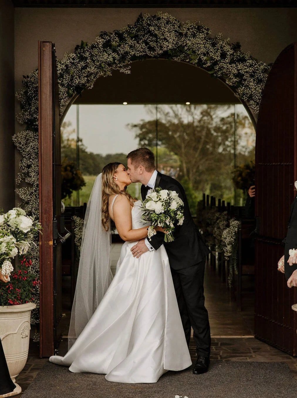 Bride and groom kissing at floral wedding arch with guests watching