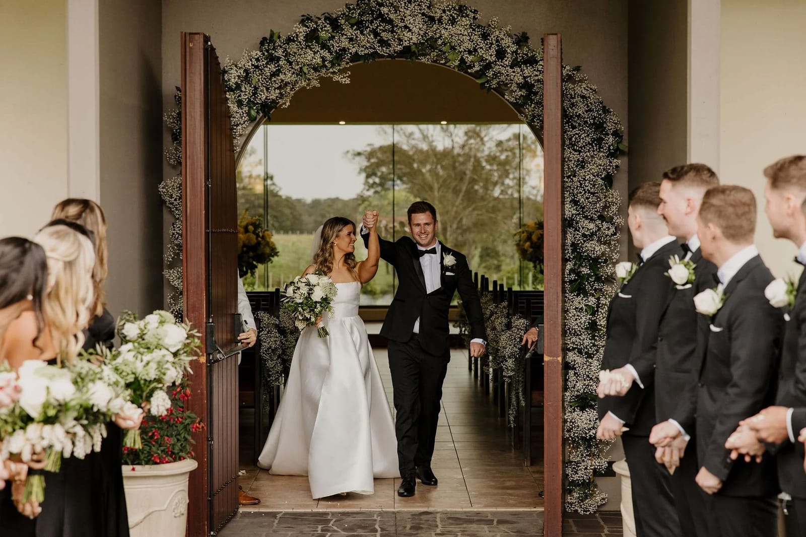 Bride and groom walking through wedding arch with vineyard view beyond