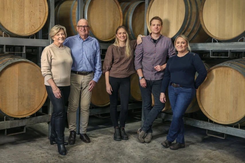 Five members of the winery team posing in front of wooden wine barrels in the barrel room