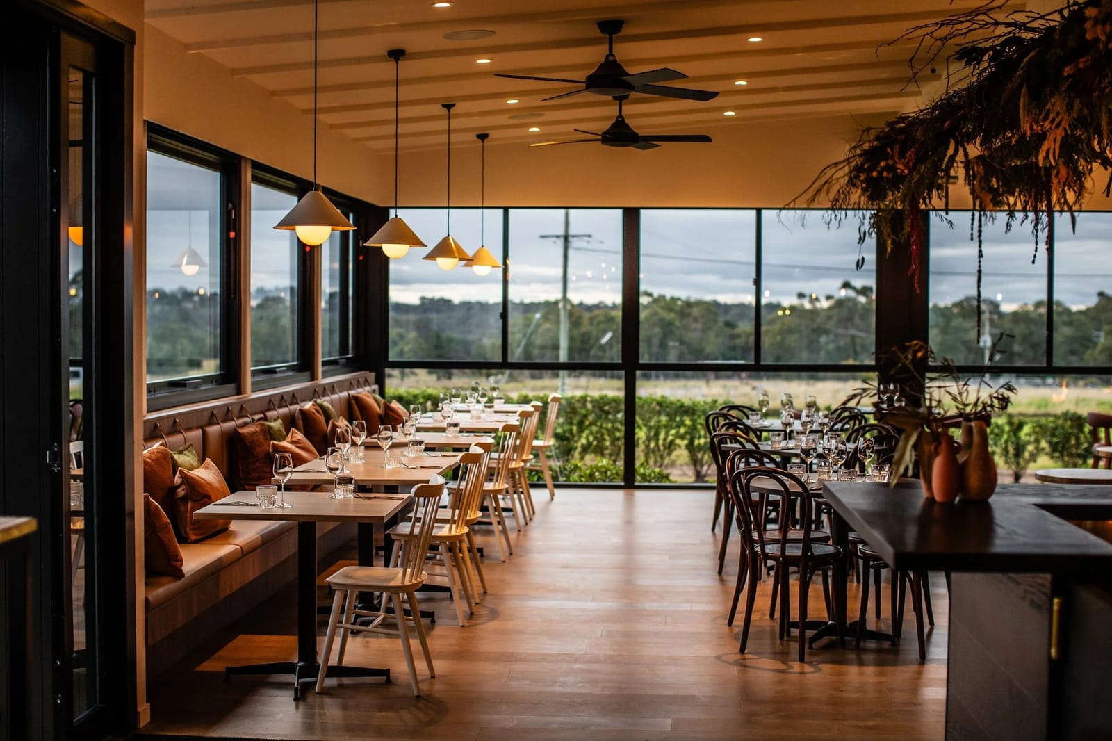 Dining room at Harkham Restaurant and Wine Bar at dusk, tables set beside floor-to-ceiling windows overlooking vineyard rows.
