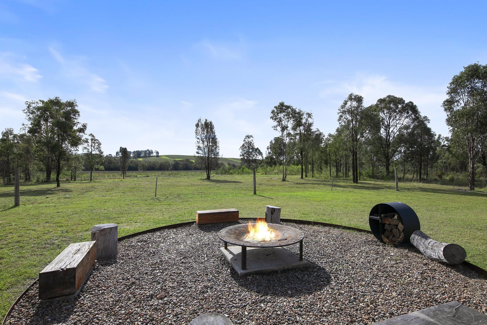 Firepit and log seating on gravel in the grounds, with cleared paddock and treeline behind.