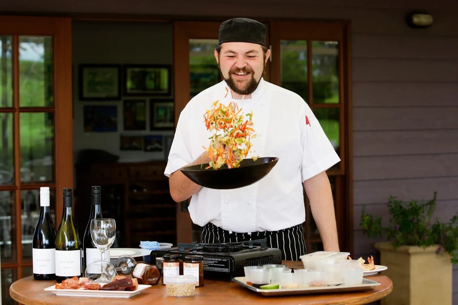 Chef Ben tossing a wok of vegetables at an outdoor cooking class bench set with bottles of local wine