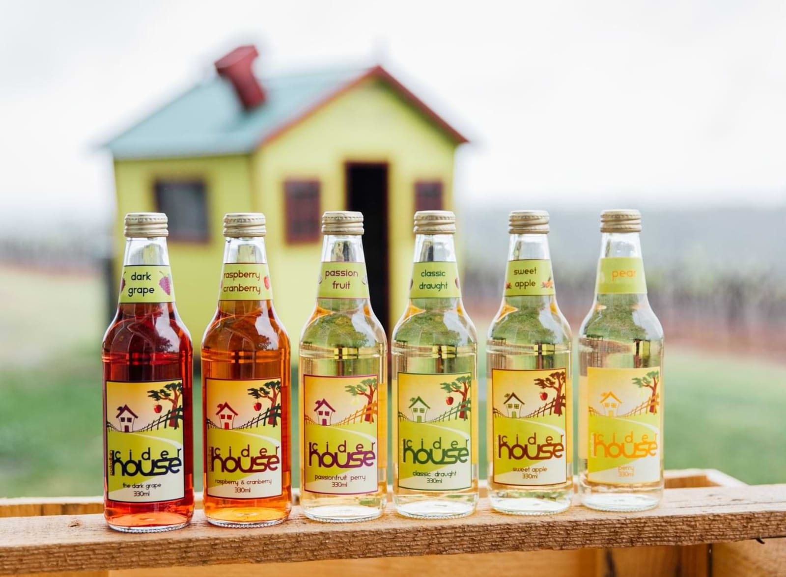 Six cider bottles lined up on a rail with the cider house behind