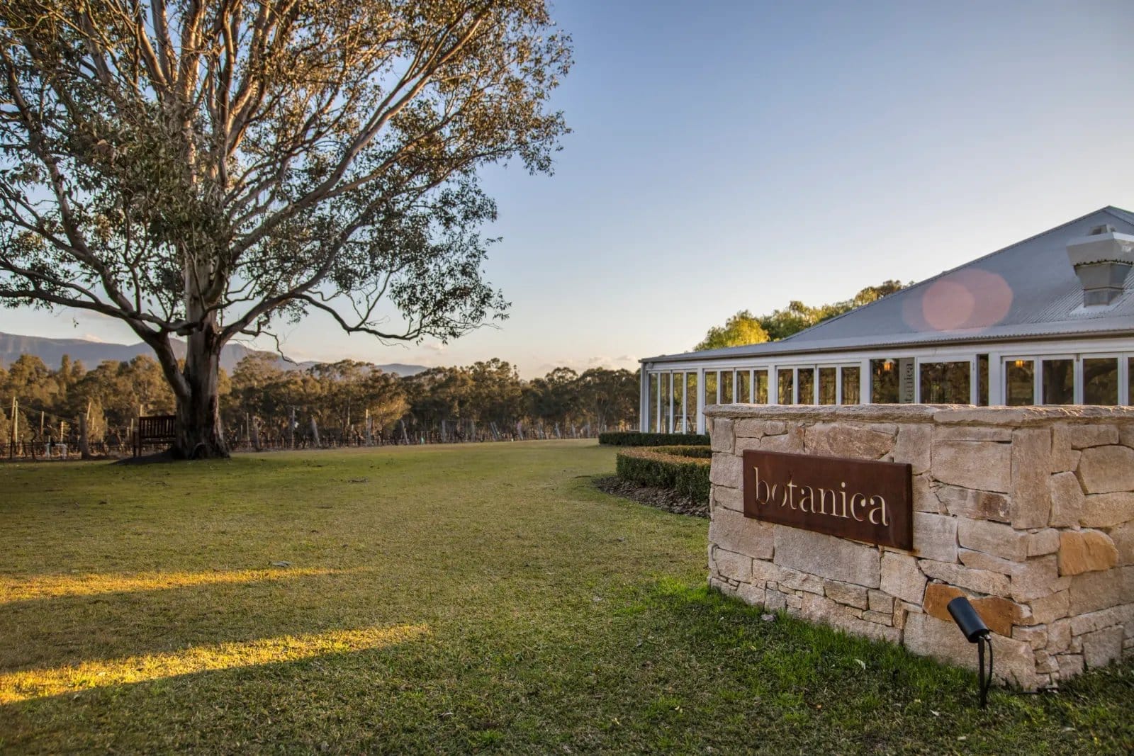The Botanica sign on a sandstone wall with the restaurant and vineyard behind at dusk