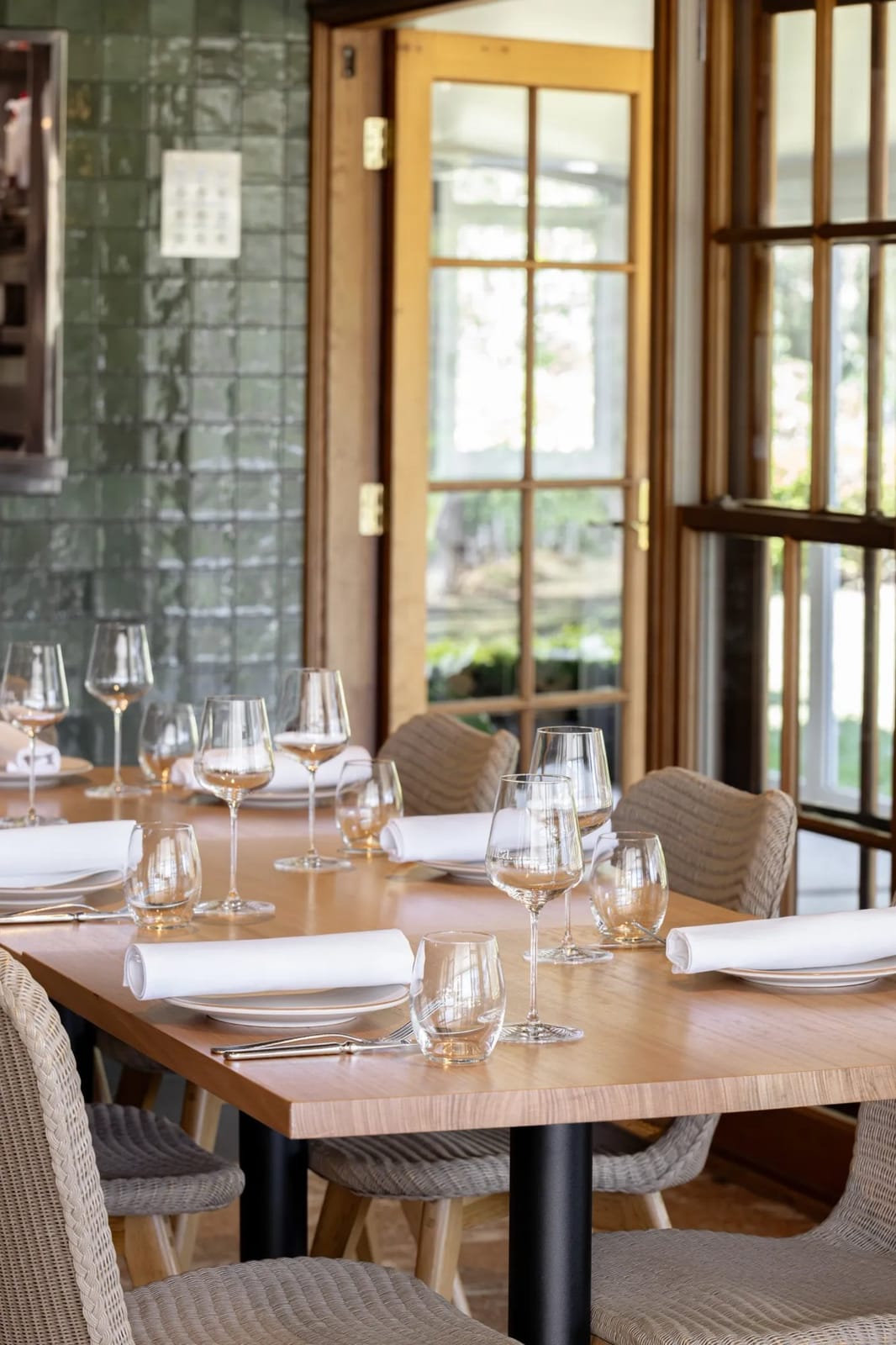 A long timber table set with wine glasses and rolled napkins beside the restaurants windows