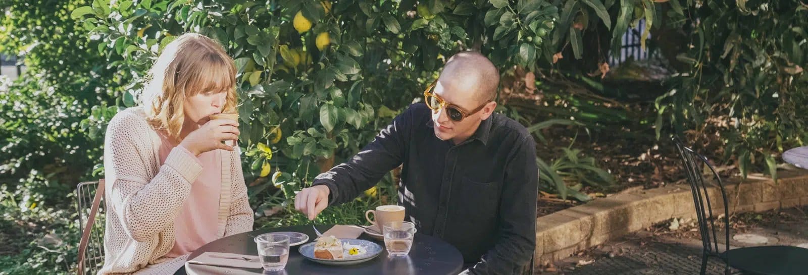 Two visitors sit at an outdoor table at Seraphine Cafe under a lemon tree, with coffee and cake.