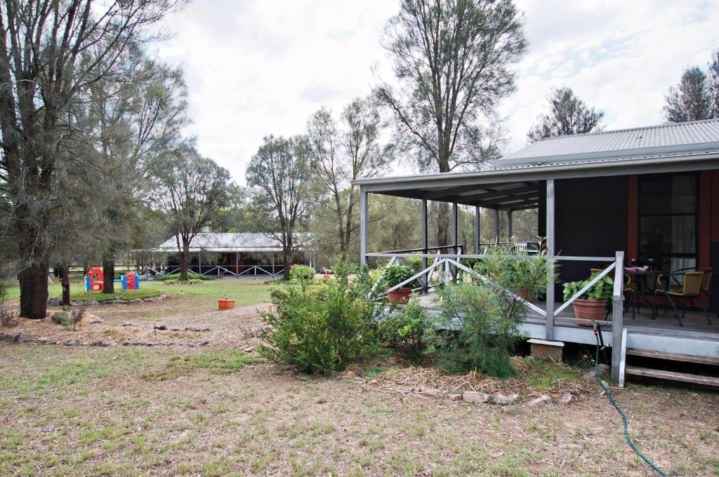 Exterior of a Serenity Grove cottage with a covered verandah and garden, a second cottage visible through the trees beyond.