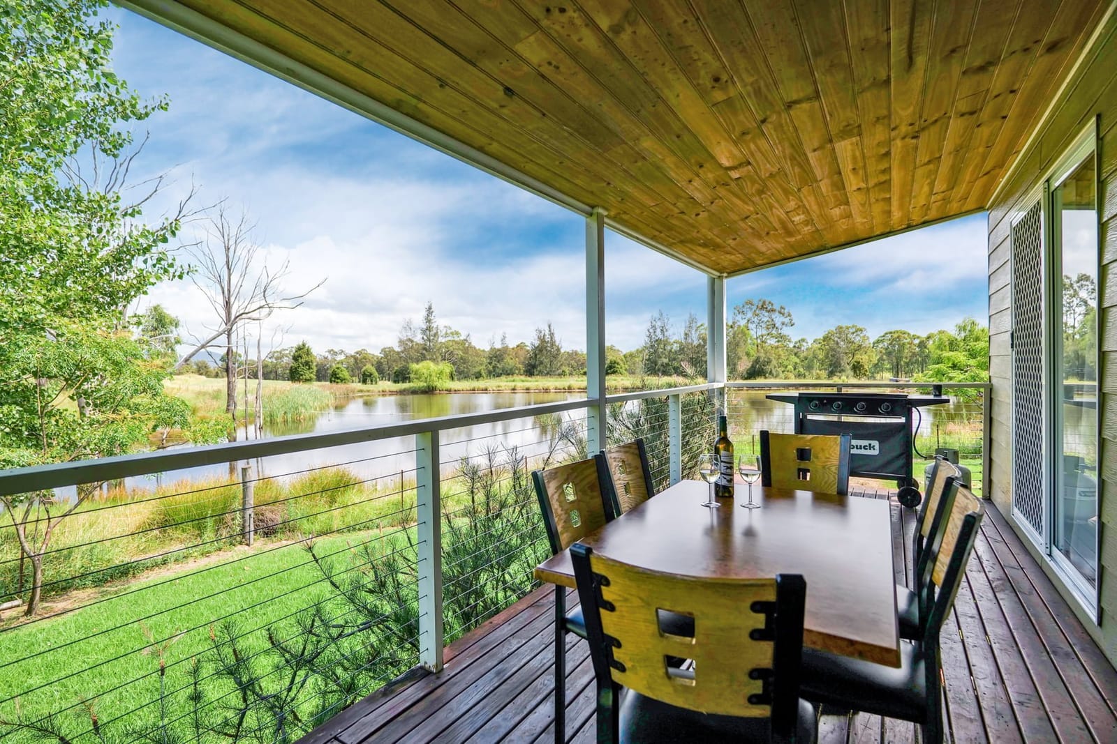 A covered cottage veranda with a barbecue and table looking over the dam