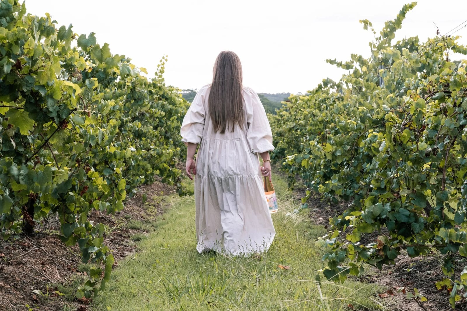 Woman in white dress walking through vineyard rows with bottle at Tatler, Lovedale, Hunter Valley