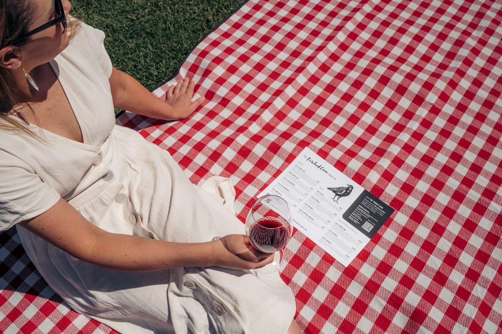 A visitor sitting on a red gingham rug on the lawn with a glass of red and the tasting list