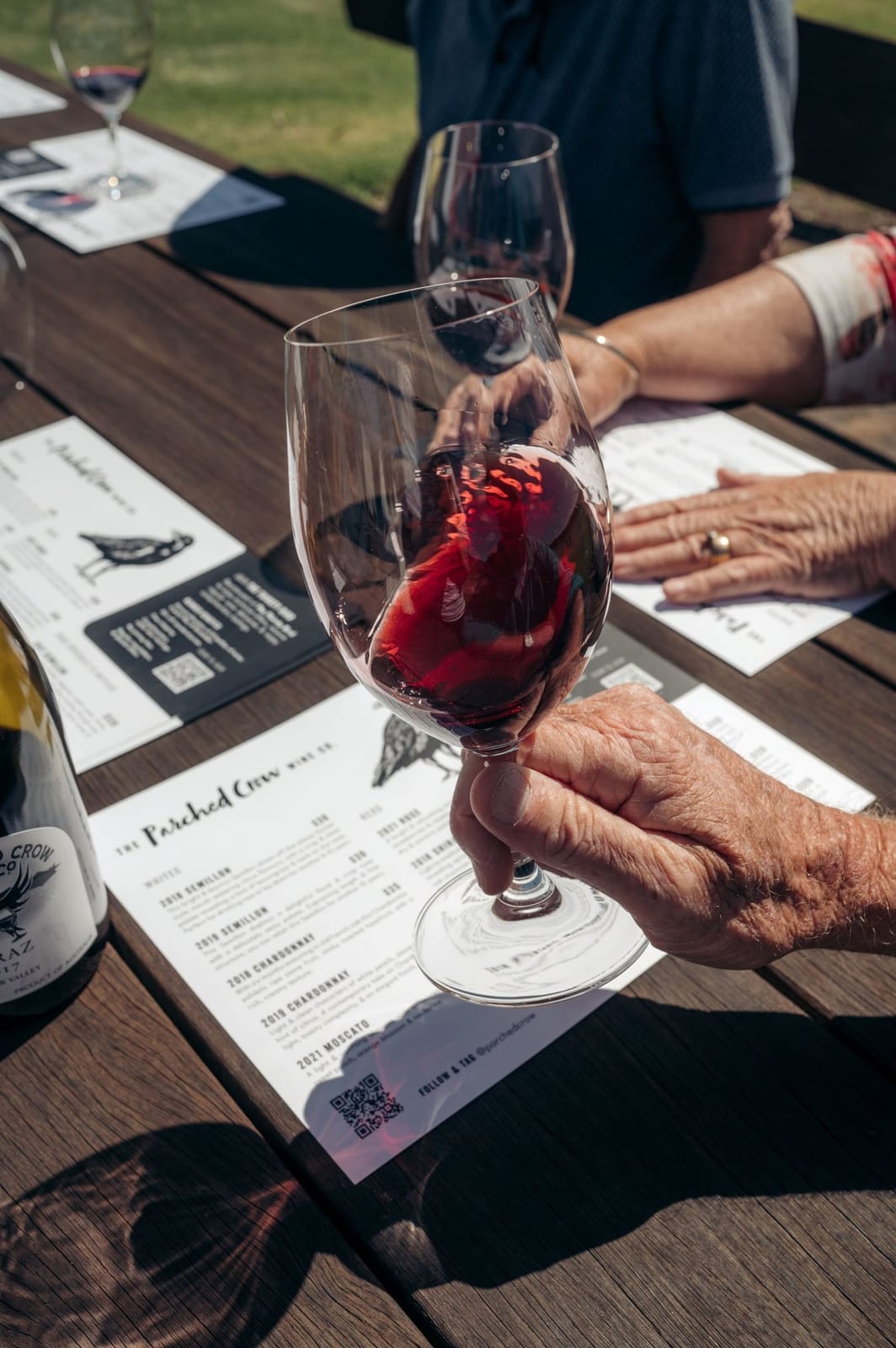 A hand swirling a glass of red over a Parched Crow tasting mat at an outdoor table