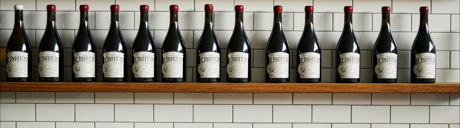 Row of Usher Tinkler wine bottles on a shelf against white tiles in the cellar door