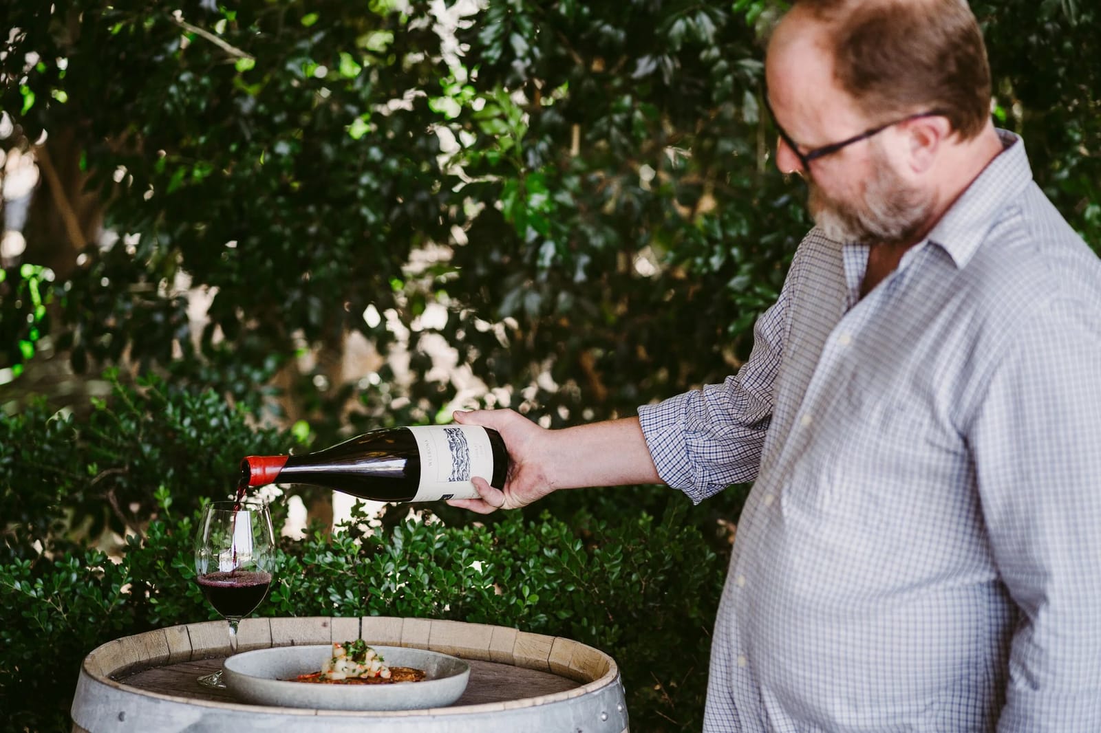 Winemaker Michael Partridge pouring a red wine next to a dish set out on a barrel head