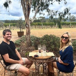 Two visitors tasting wine at outdoor table beneath shade tree